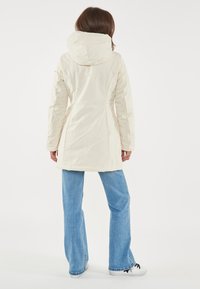 Giacca color crema, resistente all'acqua, con cappuccio, dalla forma aderente e cuciture rinforzate, abbinata a jeans azzurri svasati e sneakers.