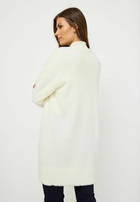 Długi, kremowy kardigan o miękkim, teksturowanym wykończeniu. Charakteryzuje się prążkowanym dołem i mankietami. Nosi się go na ciemnych dżinsach, widok z tyłu.