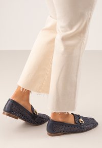 Mocasines de tejido azul marino con una suela plana marrón, que presentan una hebilla decorativa dorada y un patrón texturizado; combinados con pantalones acampanados de color claro.