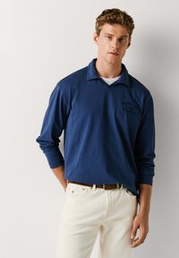 Granatowa koszulka polo z długim rękawem i kołnierzykiem; na piersi delikatne haftowane zdobienie, zestawiona z kremowymi spodniami.