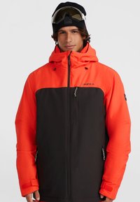 Tweedekleurige skijas in oranje en zwart. Bevat een ritsborstzak, een verstelbare capuchon en versterkte manchetten met klittenbandsluitingen.