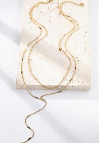 Goldfarbene lange Halskette mit mehreren geschichteten Ketten, die flache Glieder und strukturierte Akzente aufweist. Auf einem hellen Steinhintergrund präsentiert.