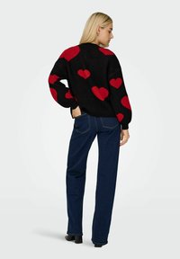 Svart tröja med röda hjärtmönster, ballongärmar och ribbad fåll, tillsammans med mörkblå raka jeans och svarta ankelstövlar.