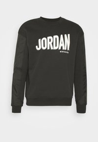 Sweat-shirt noir à manches longues et col rond avec le texte "JORDAN" en blanc sur la poitrine et des panneaux en tissu brillant sur les manches.