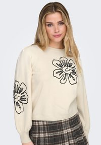 Kremfarvet sweater med sort blomsterbroderi på ærmerne, med rund halsudskæring og ribbet kant. Stylet med et ternet skørt.