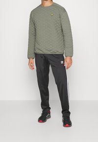 Felpa imbottita verde oliva con maniche lunghe e collo tondo, abbinata a pantaloni da jogging neri con strisce laterali bianche e un logo.