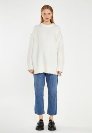 Suéter blanco de gran tamaño de punto con puños acanalados, combinado con unos vaqueros claros recortados. El modelo lleva zapatos de charol negros. Diseño sencillo y texturizado.