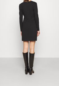 Kobieta w dopasowanej, czarnej sukience z długim rękawem i skórzanych, czarnych butach do kolan, stoi tyłem do aparatu.