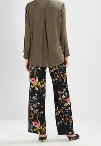 Chemise vert olive à manches longues avec pli au dos ; pantalon noir à jambes larges orné de motifs floraux colorés ; chaussures noires à talons.
