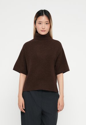 Brauner, oversized Rollkragenpullover aus weichem Strick, mit kurzen Ärmeln und lockerer Passform, kombiniert mit dunkel gestreiften Hosen.