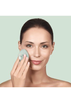 Frau mit klarer Haut verwendet eine handgehaltene Silikon-Gesichtsreinigungsbürste auf ihrer Wange vor einem blassgrünen Hintergrund.