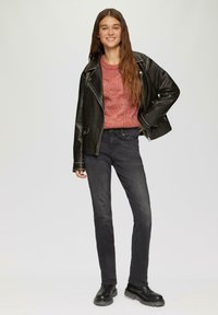 Zwarte leren motorjack met zilveren hardware, rode gebreide trui en donkere jeans. Model draagt zwarte enkellaarsjes en staat tegen een lichte achtergrond.