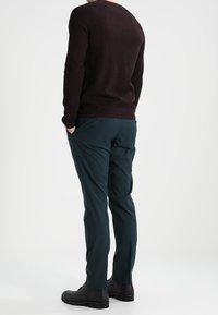 Mężczyzna stojący z rękami w kieszeniach, ubrany w ciemnobrązowy sweter, ciemnozielone spodnie i czarne eleganckie buty, na jednolitym tle.