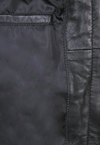 Schwarzes Ledergewebe mit glatter Textur, das über ein Taschendetail und eine Nahtlinie verfügt. Das Innere besteht aus einem matten schwarzen Material.