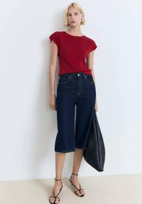 Top rosso a maniche corte abbinato a jeans blu scuri a vita alta. La modella tiene una grande borsa nera intrecciata e indossa sandali neri con lacci.