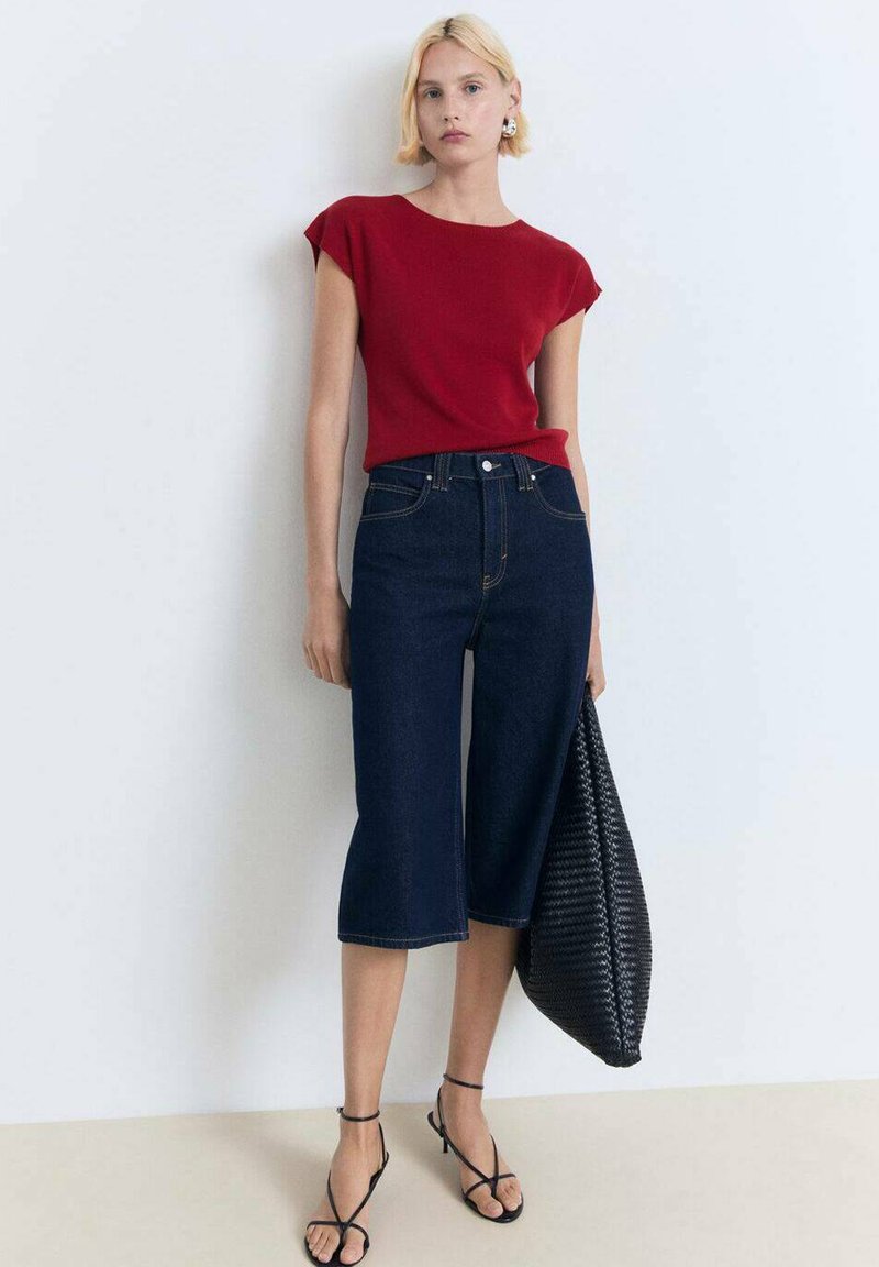 Top rosso a maniche corte abbinato a jeans blu scuri a vita alta. La modella tiene una grande borsa nera intrecciata e indossa sandali neri con lacci.