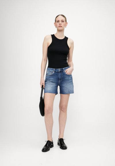 Juodas be rankovių marškinėlis, derinamas su šviesiai mėlynomis džinsinio audinio šortais, turinčiais nusidėvėjusią apdailą. Modelis laiko juodą rankinę ir dėvi blizgančius juodus batus.