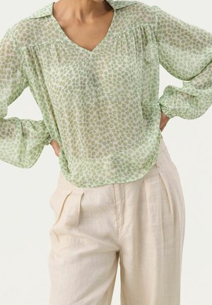 Blouse verte transparente à motif floral, col en V, manches longues avec poignets froncés, associée à un pantalon taille haute beige clair.