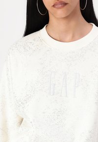 Biely sveter s textúrovaným povrchom, s jemným bodkovaným vzorom a vyšitým logom "GAP" v svetlo šedej farbe na prednej strane.