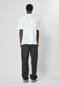 Tricou alb din bumbac cu mâneci scurte, purtat cu blugi din denim de culoare închisă. Croială lejeră, cu cusături vizibile și buzunare pe spate. Tenisi transparenți.