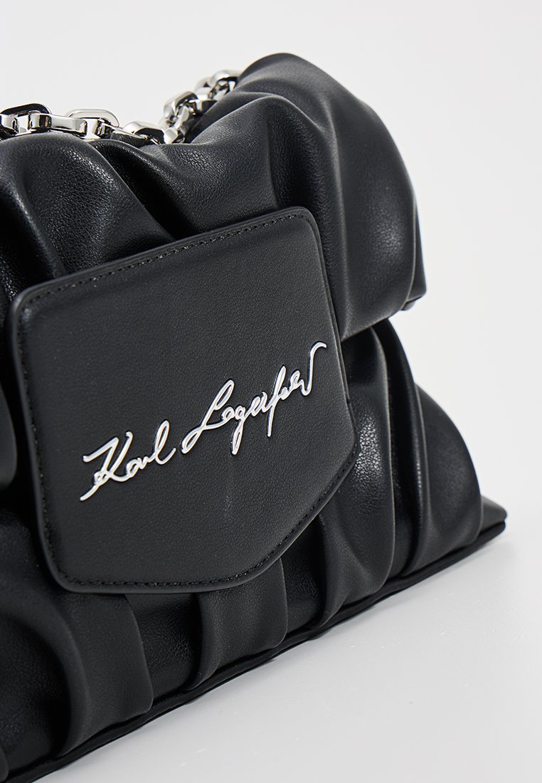 Bolso de mano negro de cuero sintético acolchado con textura suave, una correa de cadena plateada y una solapa frontal que presenta el nombre grabado "Karl Lagerfeld".