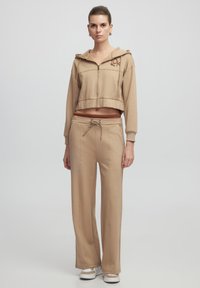 Beige cropped hoodie met een voorrits en logo, gecombineerd met beige drawstring broek met een relaxte pasvorm en rechte pijpen.