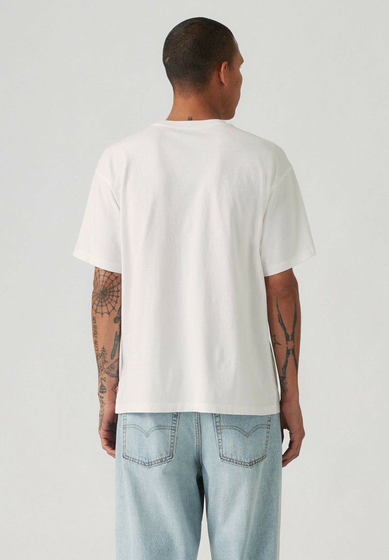 Mand med kort hår og tatoveringer iført almindelig hvid kortærmet t-shirt og lyseblå jeans, set bagfra mod en ensfarvet baggrund.