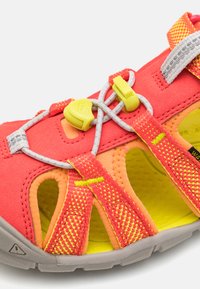 Helle korallenrote und gelbe Sportsandalette mit Netzfutter, elastischen Schnürsenkeln und strukturierten Riemen; verfügt über Aussparungen zur Belüftung.