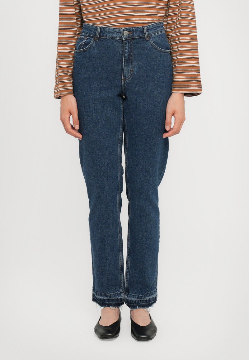 Σκούρες μπλε τζιν με ίσια πόδια, με ξεθωριασμένη υφή, ψηλ�ή μέση και ξέφτια στο τελείωμα, συμπληρωμένα από μαύρες φλατ παπούτσια.