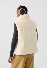 Weiße Steppweste mit hohem Kragen und strukturierter Stoffoberfläche, getragen über einem dunklen Langarmshirt. Braune gesteppte Kopfhörer werden getragen.