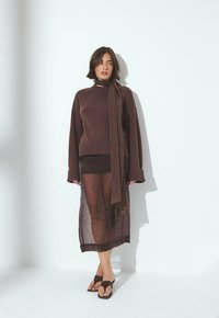 Femme portant un pull surdimensionné marron, une jupe midi transparente marron, une écharpe assortie et des sandales ouvertes marron, se tenant contre un mur blanc.