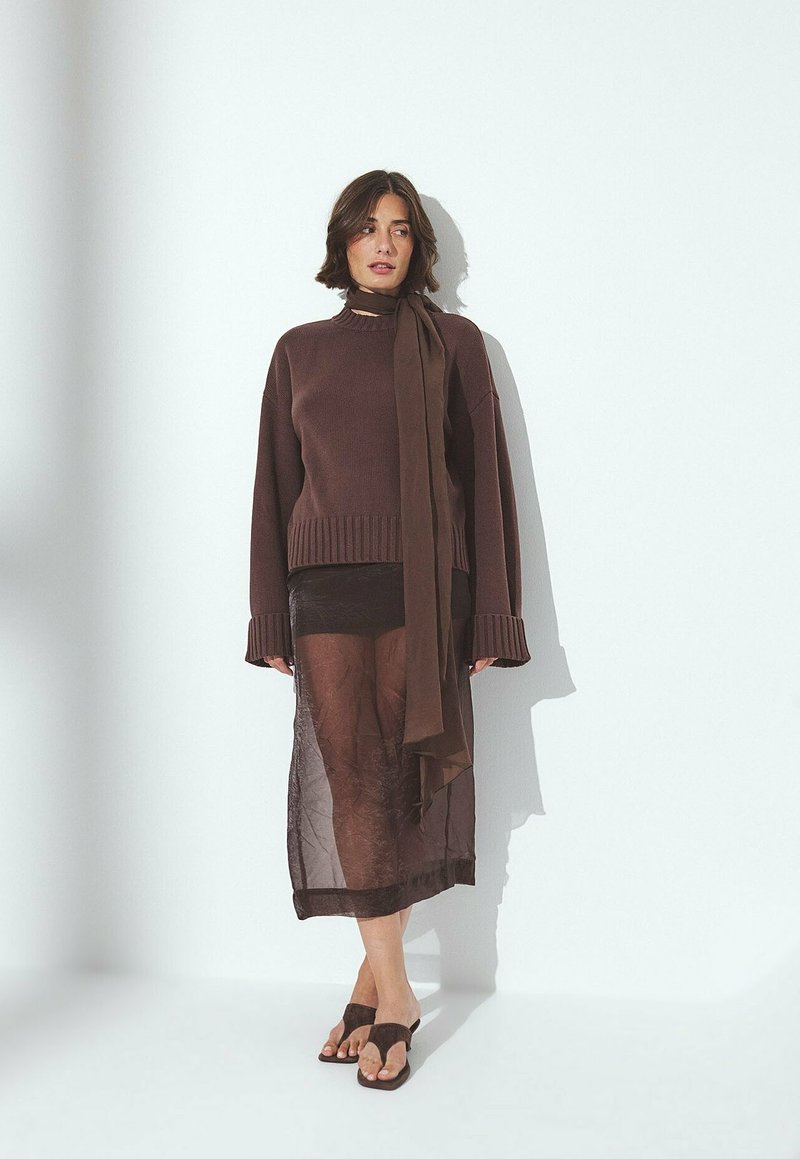 Femme portant un pull surdimensionné marron, une jupe midi transparente marron, une écharpe assortie et des sandales ouvertes marron, se tenant contre un mur blanc.