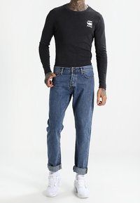 Musta pitkähihainen paito, jossa valkoinen logo, vaaleansiniset farkut ja valkoiset urheilukengät. Farkkujen hihansuut on kevyesti käännetty.