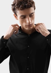 Camicia nera con bottoni e colletto, in tessuto liscio, maniche lunghe e una leggera lucentezza, con bottoni rotondi e un design aderente.