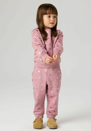 Malo dijete u ružičastoj majici s točkicama i hlačama, stoji s rukama sklopljenim, gledajući blago udesno na običnoj pozadini.