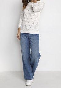 Valkoinen neulepaita, jossa on timanttikuvio, yhdistetty löysiin sinisiin farkkuihin ja valkoisiin tennareihin. Ominaisuuksiin kuuluu pyöreä kaula-aukko ja kolmannespuolihiharat.