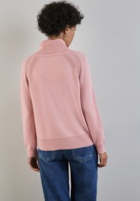 Rosa gestrickter Pullover mit hohem Kragen, gerippten Bündchen und Saum. Besitzt strukturierte Schulternähte und eine lockere Passform. Getragen mit blauen Jeans.