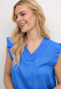 Top blu con scollo a V, maniche arricciate e smocking testurizzato lungo il collo. Tessuto liscio con una finitura di colore solido.