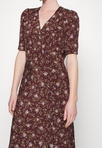 Femme portant une robe bordeaux à fleurs avec des manches courtes bouffantes et un décolleté en V, ornée de petites fleurs bleues, blanches et pêche.