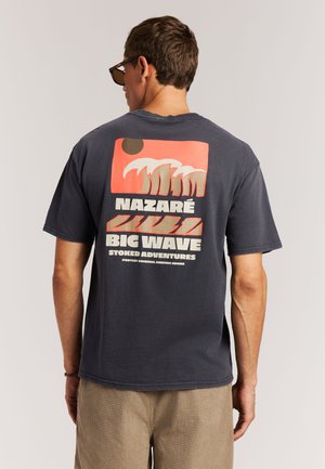 Marineblauw T-shirt met een grafisch ontwerp in roze en crème, met golven, zon en de tekst: "Nazare Big Wave Stoked Adventures." Korte mouwen.