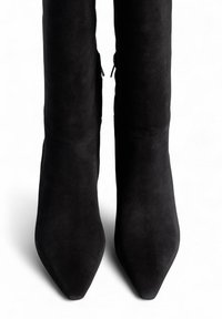 Une paire de bottes noires en daim montantes, à bout pointu et avec des zips latéraux, sur un fond blanc.