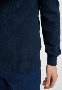 Tengerészkék, texturált kötött pulóver, bordás alsó szegéllyel és mandzsettával, oldalán címkés részlettel. Sötétkék farmernadrággal viselve.