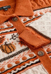 Cardigan lavorato a maglia con motivo arancione e crema, caratterizzato da una texture a treccia, con bottoni marroni e motivi decorativi, tra cui zucche e righe.