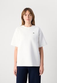 White cotton t-shirt with a relaxed fit, short sleeves, and a small green Lacoste logo on the left chest. Smooth texture.