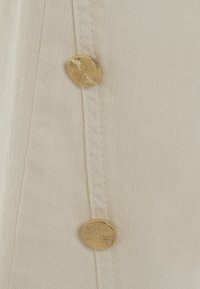 Tessuto di colore crema con una finitura testurizzata, caratterizzato da due prominenti bottoni dorati con un design a raggi solari lungo il bordo.