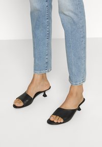 Mules de cuero negro con punta abierta, una amplia correa superior y un tacón cónico. Combinados con unos jeans rectos de color azul claro.