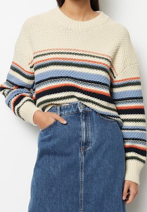 Personne portant un pull en maille épaisse avec des rayures horizontales multicolores et un jean bleu à taille haute, main dans la poche.