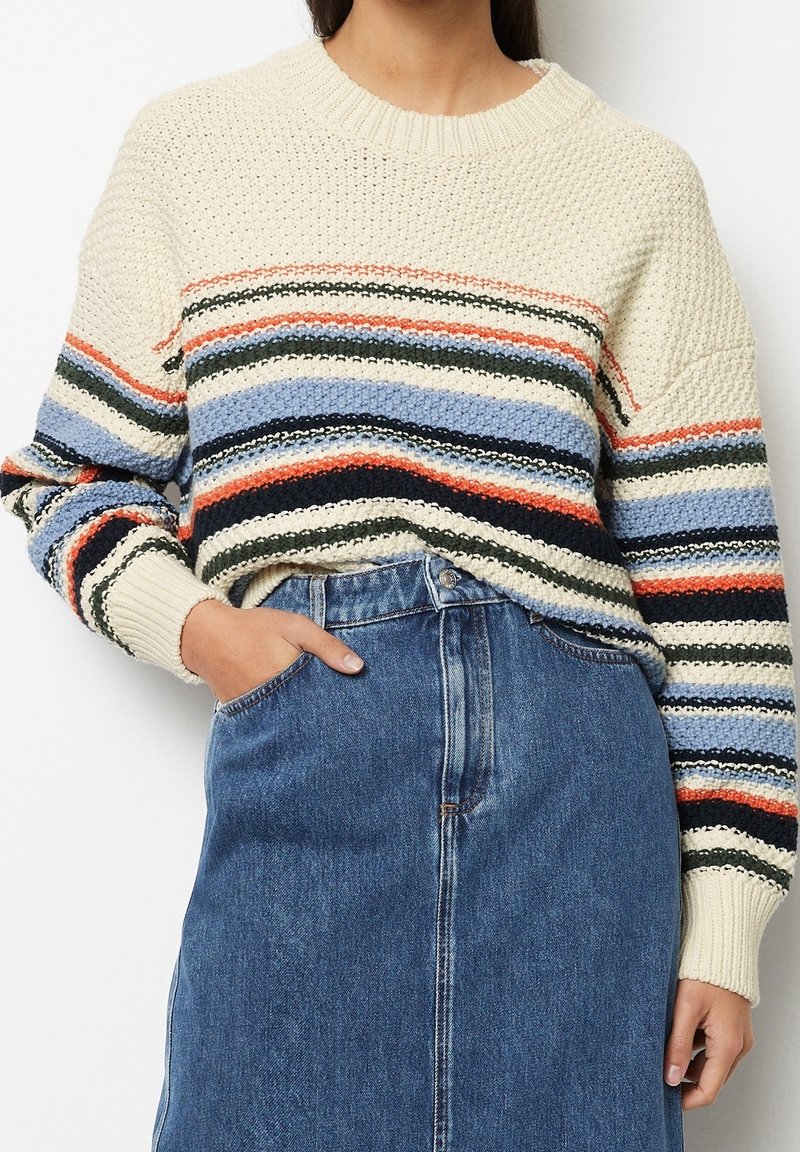 Personne portant un pull en maille épaisse avec des rayures horizontales multicolores et un jean bleu à taille haute, main dans la poche.