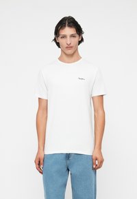 Tricou alb din bumbac cu guler rotund, mânecă scurtă și un logo brodat negru pe pieptul stâng. Purtați cu blugi de culoare albastră deschisă.
