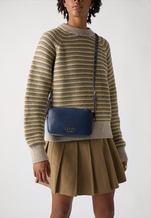 Borsa a tracolla blu in pelle con logo impresso, tracolla corta, forma rettangolare, texture liscia, indossata con un maglione a righe e una gonna plissettata.